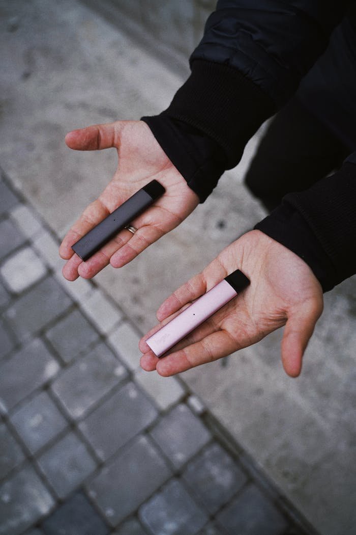 services-03 Close-up of hands holding two colorful vape pens on a city pavement.
