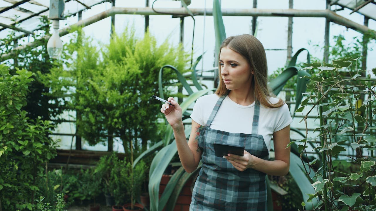 services-01 Woman in checkered apron observing plants in a greenhouse setting.