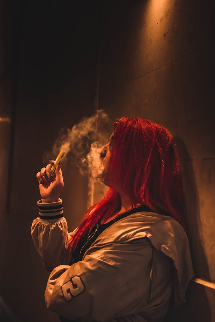 gallery-2 Dramatic indoor shot of a woman with red hair vaping against a wall.